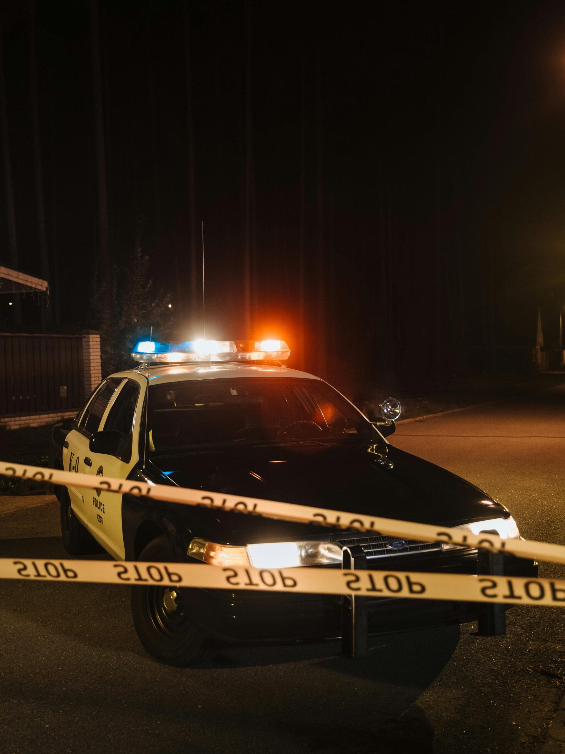 A police car with emergency lights at a night crime scene, surrounded by yellow police tape.